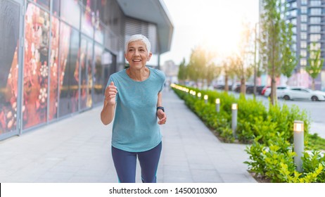 Front View Of Senior Woman Jogging Through Park. Elderly woman running with smartwatches in the park in evening sunset. Attractive looking mature woman keeping fit and healthy. - Powered by Shutterstock