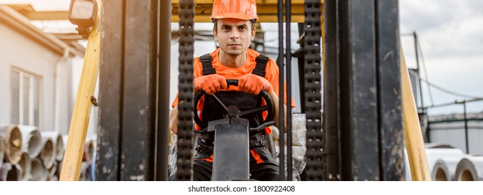 Front view of a serious face young caucasian man builder in overalls and orange protective helmet drive a construction loader machine on the construction materials store
