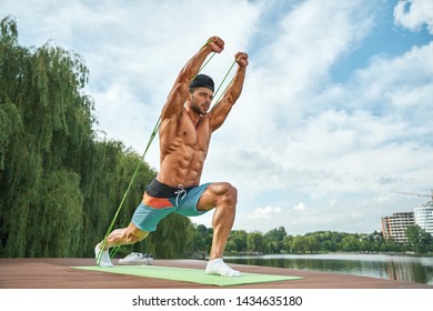Front view of serious male stretching with silicons expander, pulling hands up. Athletic man with bare chest and sunburn doing lunge exercise and enjoying of beautiful landscapes.