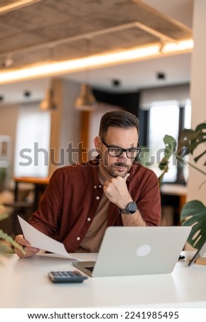 Front view of serious millennial man using laptop sitting at the working places, focused guy communicating online, writing emails, distantly working or studying on computer.