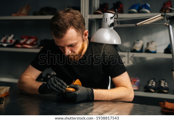 Front view of shoemaker wearing black latex gloves\
rubbing old light brown leather shoes for later restoration.\
Concept of cobbler artisan repairing and restoration work in shoe\
repair shop.