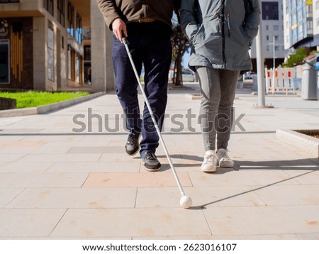 Front view of a sightless man walking on the pavement using his cane and a woman providing guidance.