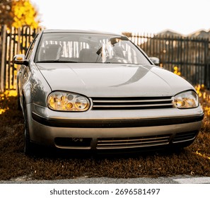 Front view of a silver or gray compact hatchback car, illuminated by warm, golden sunlight. The vehicle is parked outdoors in a residential area, with a wooden fence and autumnal foliage in the soft-f