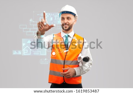 Front view of smiling architect with paper rolls wearing reflective vest and helmet. Digital tactile charts screen, man touching virtual icon on projection. Concept of digitalization, construction.