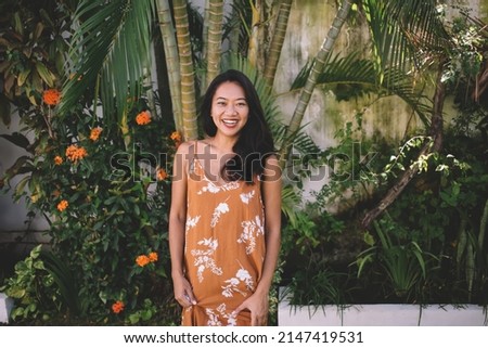 Front view of smiling asian girl posing in yard with tropical plants on Bali island. Cropped image of young beautiful brunette indonesian woman in dress looking at camera at sunny day