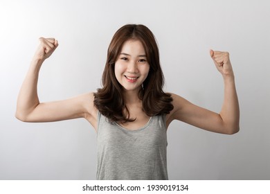 Front view of A smiling beauty young Asian girl indoor photo of young pretty lady dressed in basic gray t-shirt smiling broadly at camera and demonstrating her strong bicepses, over white background.