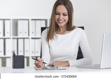 Front view of a smiling blond woman wearing white clothes and writing in her clipboard while sitting at her workplace.