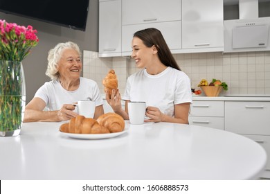 Front view of smiling relatives drinking tea and spending time together in white kitchen. Young brunette laughing and showing croissant to happy grandmother. Concept of family. - Powered by Shutterstock