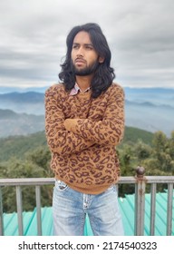 Front View Of A South Asian Young Guy With Long Hair And Beard Looking Sideways While Posing With Crossed Arms, Standing With Leaning On Safety Barrier Against The Background Of Mountains