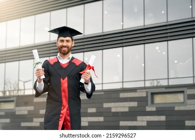 Front View Of Student With Master Degree Standing, Holding Two Diplomas, Smiling. Handsome Male In Mortarboard And Graduate Gown Graduating From High School. Concept Of Youth.