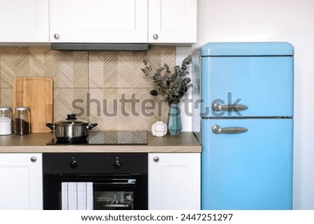 Front view of stylish interior design in kitchen. White furniture, tiled wall, built-in appliances, blue retro refrigerator in apartment. Kitchenware and home decor on wooden countertop