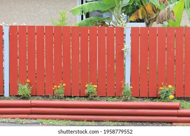 Front View Of A Stylish Residential Fence. Orange Rust Painted Wood Slats, Grey Poles, Flower Beds With Small Yellow Flowers, Horizontal Logs Positioned In Flower Boxes