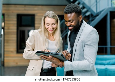 Front view of successful smiling young multiracial business people, Caucasian woman and African bearded man, standing in the office room and talking each other, looking at tablet pc