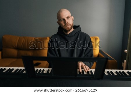 Front view of talented bald composer male with beard playing electric piano or digital synthesizer, practicing and composing music, creating melodic compositions in home living room.