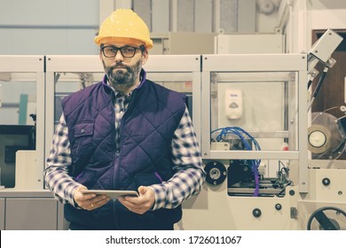 Front View Of Technician Holding Tablet At Plant. Confident Factory Worker Using Tablet While Standing Near Printing Machine And Looking At Camera. Print Manufacturing Concept