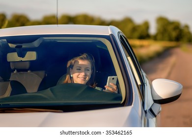 Front View Of Teenage Girl Holding Cell Phone While Driving Car On Country Road - Distracted Teenage Driver Texting Behind Wheel In White Car