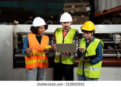 Front View Of Three Factory Workers, Asian Man And Woman, And African Woman, In A Safety Vest And Helmet, Standing In Front Of Industrial Machines Looking At Data In A Laptop Held In Hand At Factory.