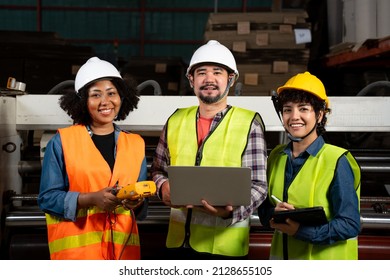 Front View Of Three Factory Workers, African Woman, Asian Man And Woman, In A Vest And Helmet, Standing Smiling At Camera While Working With A Multimeter, Laptop, And Tablet Held In Hand At A Factory.