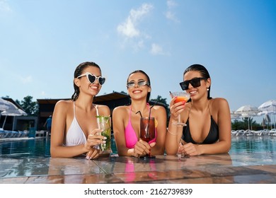 Front view of three pretty women relaxing in swimming pool. Seductive girl standing in water, holding cocktails, talking, smiling, enjoying. Concept of vacation and youth.