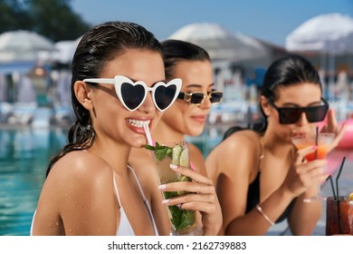 Front view of three seductive girls relaxing in swimming pool outdoors. Slim pretty women standing in water, smiling, talking, drinking cocktails. Concept of rest and summer.