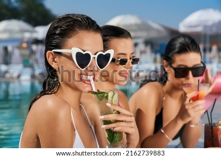 Front view of three seductive girls relaxing in swimming pool outdoors. Slim pretty women standing in water, smiling, talking, drinking cocktails. Concept of rest and summer.