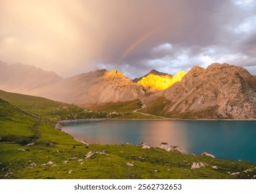 The front view of Tientang Hu lake, Xinjiang, China. There are many mountains behind. The sun was setting with soft yellow light. There is a lush green lawn and a rainbow with mist on a cloudy day. - Powered by Shutterstock