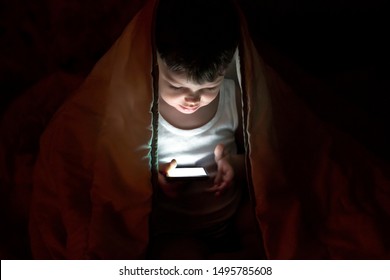 Front View Of Toddler With Face Lit From Phone Screen At Night. Image Of Nice Boy In Pajama Looking At Mobile. Portrait Of Charming Addicted To Phone Adorable Boy,holding Smartphone In His Hands. 