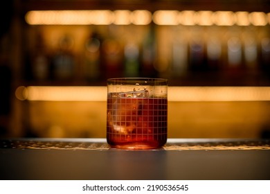 Front View Of Transparent Glass With Grid Pattern Full Of Cold Cocktail On Bar Counter. Blurred Bar Background