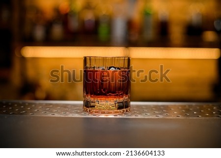 front view of a transparent old-fashioned glass with alcoholic drink and a piece of ice on a bar counter against a blurred background