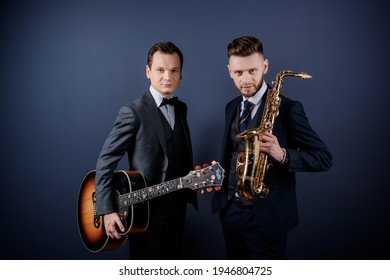 Front view of two men holding musical instruments guitar and saxophone looking at the camera. Men standing on the blue wall isolated.