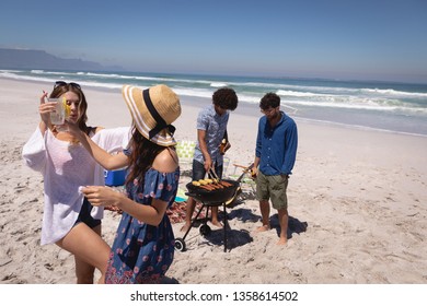 Front view of two Multi ethnic women friends dancing while men friends doing a barbecue with ocean waves in background