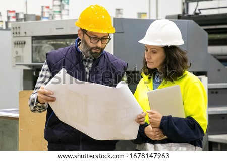 Front view of two printing workers inspecting quality of print. Focused employees looking at blueprint. Print manufacturing concept