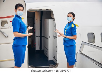 Front View Of Two Slim Female Flight Attendants Standing At The Entrance To The Airplane