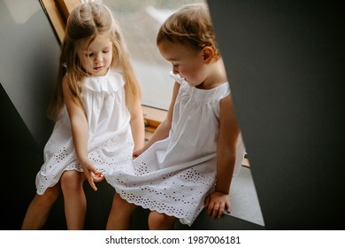 Front view  of  two small adorable girls sitting on the window-sill. Sisters dressed in white dresses.