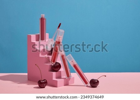 Front view of unbranded lipsticks decorated with fresh red ripe cherries on pink stair on a blue background. Cherry-based lip balm helps keep lips fresh and rosy. Advertising photo and copy space
