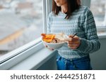 Front view unrecognizable woman standing at the window at home holding a plate and a spoon with a mixture of cereals with raw fruits. A young girl eats right and maintains a good mood and health.