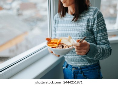 Front view unrecognizable woman standing at the window at home holding a plate and a spoon with a mixture of cereals with raw fruits. A young girl eats right and maintains a good mood and health. - Powered by Shutterstock