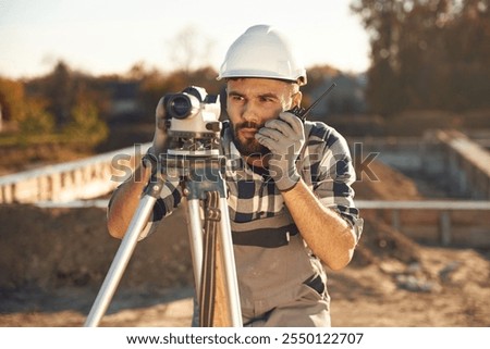 Front view, using geodetic optical level. Worker is on the construction site at daytime.