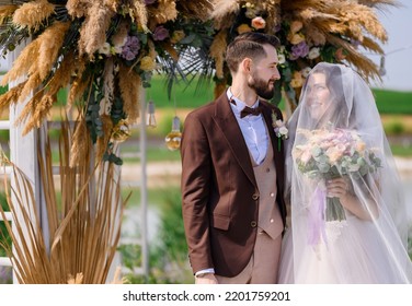 Front view of wedding ceremony of groom in brown tuxedo and bride with wedding veil on face wearing in puffy dress, looking at each other while standing near decorated arch with dried flowers - Shutterstock ID 2201759201