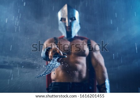 Front view of wet roman gladiator in iron helmet and red cloak pointing sword at camera. Selective focus of raindrops on blade in arms of muscular shirtless spartan in armor in rainy bad weather.
