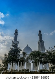The front view of the white mosque with two minarets is with several trees in the yard and a gradient blue sky in the background 