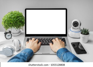 Front view of woman hands working on laptop with blank white screen standing on the white office desk with smartphone and web- camera. Mock up, copy space for your text.