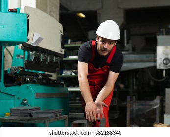 front view of a worker, wearing red overalls, and a white protective helmet, bending over to pick a bent piece of metal, next to an industrial bending machine, painted in turquoise and white