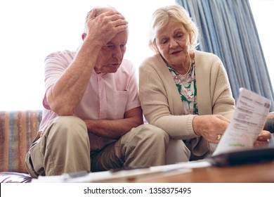 Front View Of Worried Senior Caucasian Couple Discussing Over Medical Bill While Sitting On Sofa At Retirement Home. Senior Male Has Hand On His Head.