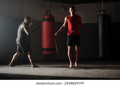 Front view of young active man with skipping rope jumping on the floor of gym or sports center with guy practicing kicks on background - Powered by Shutterstock