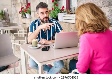Front view of a young adult with a beard and glasses talking about work with a blonde woman sitting on their backs while having a coffee. Two adults chatting amicably on the bar' terrace. - Shutterstock ID 2196704035
