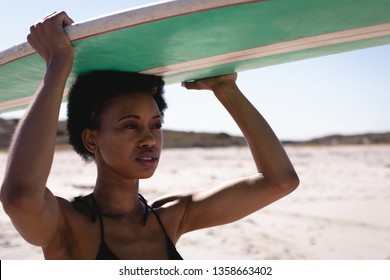 Front view of young African American woman carrying her surfboard on her head at beach in the sunshine