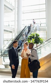 Front View Of A Young African American Businessman Talking With A Young Caucasian Businessman And Businesswoman In The Atrium Of A Busy Modern Building. Modern Corporate Start Up New Business Concept