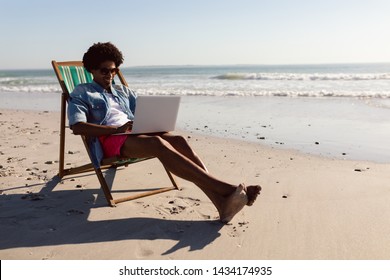 Front View Of Young African-american Man Using Laptop While Relaxing In A Beach Chair On The Beach