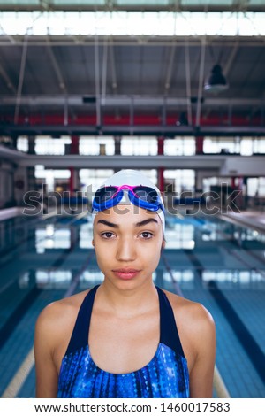 Front view of a young African-American woman wearing a swimsuit and swimming cap with goggles looking at the camera while standing by an olympic sized pool inside a stadium